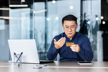 Fototapeta premium Upset and sad asian businessman inside office, man holding smartphone and bank credit card in hands, no funds in account received payment rejection, man working with laptop.