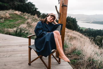 thoughtful happy blue-eyed caucasian girl with a faraway look in a brown bathrobe and cap sitting on the left back of a comfortable folding chair calm and relaxed barefoot with her hands on her - obrazy, fototapety, plakaty