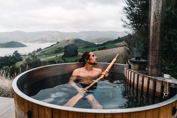 calm caucasian boy without clothes taking a bath with a long wooden spade in his hands inside a metallic hot water jacuzzi relaxing in the mountains far from civilization near the forest trees, te