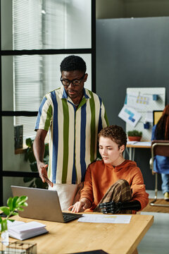 Young confident businessman in striped casual shirt explaining data to colleague while pointing at laptop screen during discussion