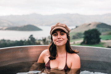 blue-eyed caucasian girl wearing a swimsuit and a hat sitting inside the nice hot water jacuzzi looking at the camera smiling calm and happy in a natural and silence environment, te wepu pods akaroa - obrazy, fototapety, plakaty