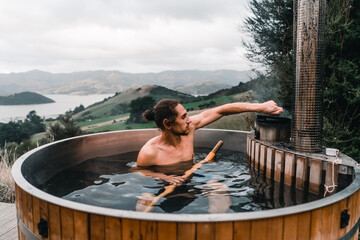 calm caucasian boy without clothes inside metallic hot water jacuzzi relaxing adding coal to boiler while bathing in mountains far from civilization near forest trees, te wepu pods akaroa, new zealand - obrazy, fototapety, plakaty