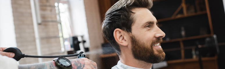 tattooed hairdresser spraying perfume on pleased bearded man in barbershop, banner.