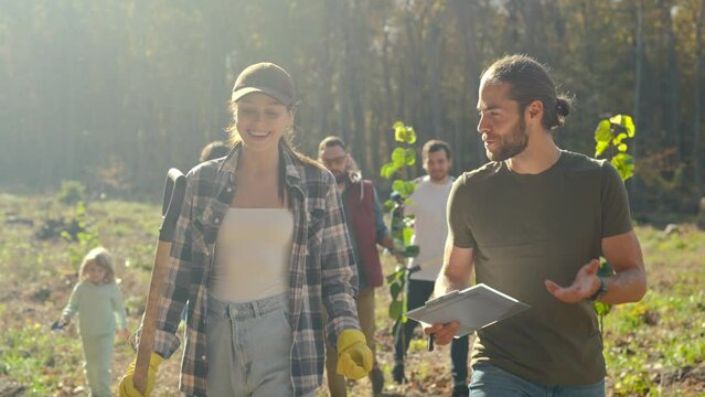 Caucasian young couple of environmental volunteers walking outdoor with other people on background, watching on some documents or plan and discussing planting trees.