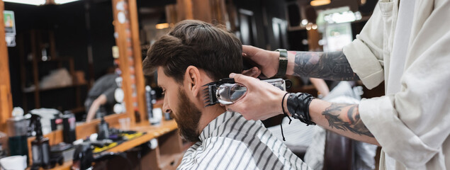 Fototapeta na wymiar Tattooed hairstylist holding trimmer near bearded client in barbershop, banner.