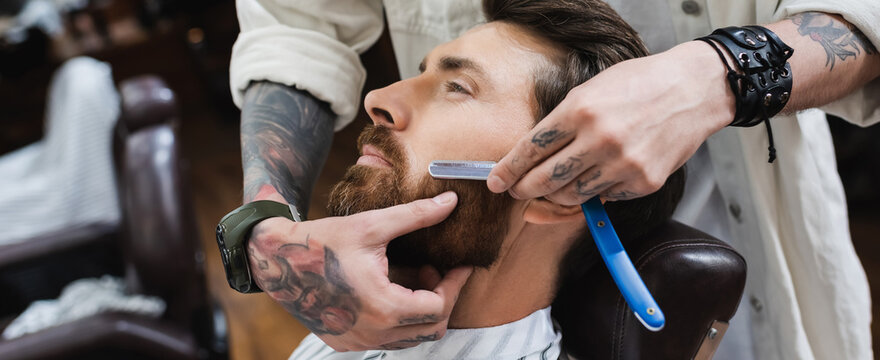 tattooed hairstylist in leather bracelet shaving man with straight razor, banner.