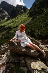 Naklejka na ściany i meble Elegant sexy lady with red hair in a white dress sits on the stone in the mountains and relaxes under sun rays. Fashion natural portrait as wallpaper, magazine design or advertising cover.