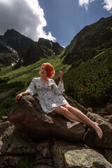 Elegant sexy lady with red hair in a white dress sits on the rock among spruce in the mountains and relaxes under sun rays. Fashion natural portrait as wallpaper, magazine design or advertising cover.