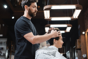 Side view of barber styling hair of teenager in cape in barbershop.