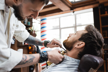 brunette bearded man near hairstylist working with comb and hair clipper.