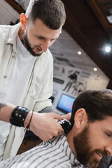 Fototapeta premium Bearded barber trimming hair of blurred customer in barbershop.