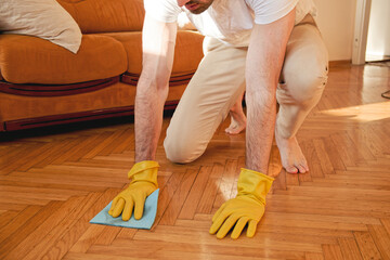 Handsome man cleaning floor in his house with a piece of cloth and using gloves, working hard - obrazy, fototapety, plakaty
