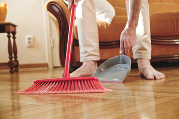 Close up of handsome man in the house, he cleaning his floor with broomstick and dustpan - obrazy, fototapety, plakaty