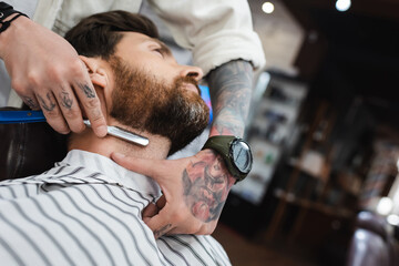 low angle view of tattooed barber in wristwatch shaving neck of bearded man with straight razor.