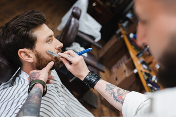 blurred barber in leather bracelet shaving client with straight razor.