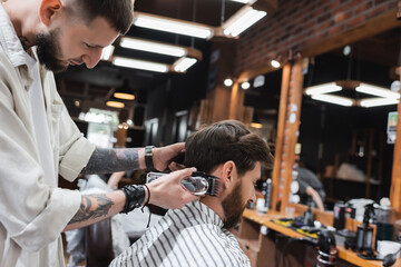 Side view of tattooed barber holding trimmer near client in barbershop.