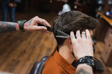 tattooed barber cutting hair of brunette man with thinning scissors.
