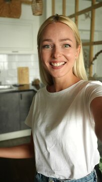 Vertical POV of pretty friendly young woman with blonde hair greeting waving hands and talking making video call, holding in hand smartphone, looking at camera, standing at home kitchen room.