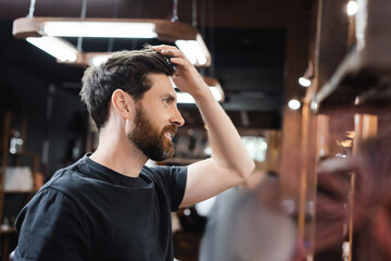 Obraz premium side view of happy brunette man touching hair in barbershop.