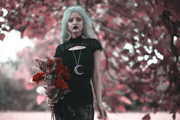 Dark goth girl standing in the forest, holding a bouquet of red roses, posing among the trees and...