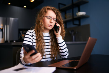 Curly blonde woman in glasses working from home with multiple electronic internet devices. Freelancer businesswoman holds tablet, talking on cellphone, has laptop on table. Multitasking theme.
