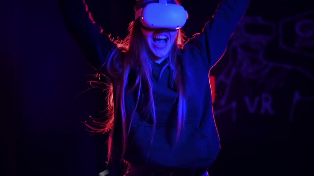 A young woman is happy about winning the VR game using VR glasses and controllers. Red and blue illumination. Slow motion