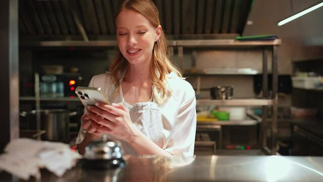 Smiling woman chef typing on mobile in the restaurant kitchen