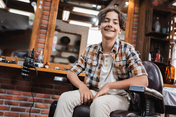 Fototapeta na wymiar Positive teenage boy in casual clothes looking at camera in barbershop.