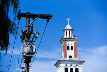 Fototapeta na wymiar Electric Transformer With Cables and Bell Tower of a Church in Blue Sky
