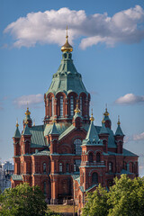 Fototapeta na wymiar Helsinki, Finland - July 20, 2022: Uspenski Cathedral. Closeup of the red brick and green domed building above green foliage under blue cloudscape