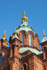Fototapeta na wymiar Helsinki, Finland - July 20, 2022: Uspenski Cathedral. Closeup of red brick and green dome main central tower with golden cross on top against blue sky. 3 minor towers included