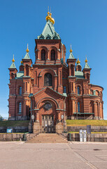 Helsinki, Finland - July 20, 2022: Uspenski Cathedral. South red brick facade with multiple towers all topped by golden pinnacle against blue sky, as seen from parking. 