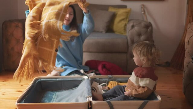 Getting ready for road trip travelling, happy family concept. Asian mother and her toddler son preparing for their travel adventure, packing clothes into suitcase. Mom and a kid pack luggage together.