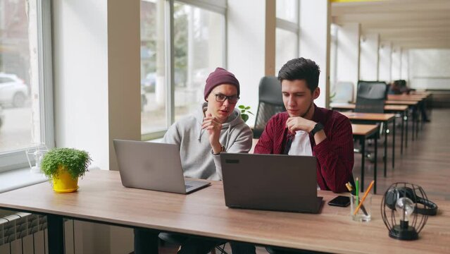 Two hipster coworkers sharing ideas about project sitting with laptops in spacious open space office. Developer discuss software program code. Designer and manager consulting on working plan.