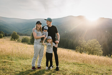 Portrait of happy young family spending time together in field. Dad, mom holds baby son and daughter stand on grass in mountains. The concept of family holiday. Mother, father hug kids on autumn day.