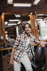Fototapeta na wymiar Positive teen boy looking at camera near armchair in blurred barbershop.