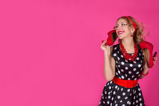 A blonde girl in a black dress with white polka dots with red accessories stands on a pink background holding high-heeled shoes in her hands. Pin-up