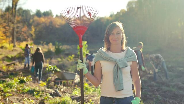 Portrait of beautiful Caucasian young woman with rake standing at forest field and smiling cheerfully to camera. Outdoors. Pretty female, eco volunteer at planting trees day. Gardening concept.