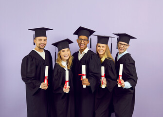 Portrait of group of happy graduates in academic mantles holding scrolls of diplomas in their hands. Proud and smiling multiracial students standing in row on purple studio background. Banner.