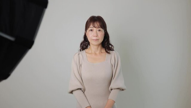 Middle aged asian woman stand still and stars the camera at photo studio. White wall.