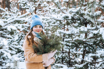 Portrait of cute caucasian teen girl with loose long hair wearing warm clothes holding green pine branches walking in snowy winter forest