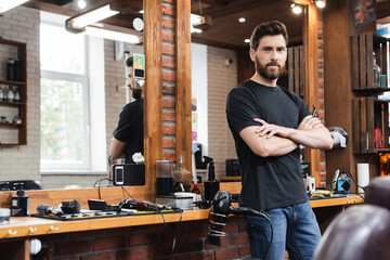 confident hairdresser standing with crossed arms and looking at camera at workplace in barbershop.