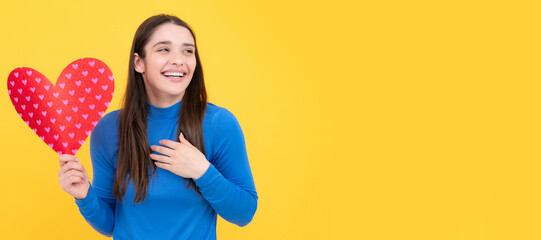Girl feeling love. Beautiful girl holding valentines gift on yellow background. Portrait of young woman holding red paper heart. Woman isolated face portrait, banner with copy space.