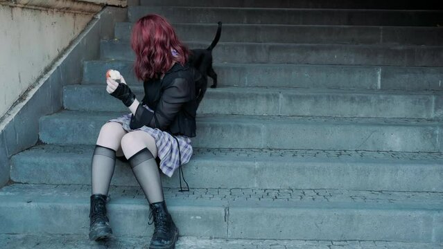 Young woman model wearing anime style clothing, red hair, sitting outdoors with street black cats 
