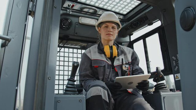 Portrait of Caucasian female technician in hard hat and coverall posing for camera sitting at driver seat of tractor cab