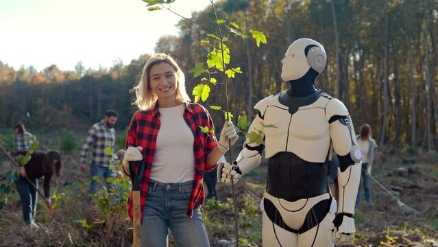 Portrait of beautiful Caucasian young woman and robot with tree seedling standing at forest field and smiling to camera. Outdoors. Pretty female, eco volunteer android for saving ecology. Gardening.