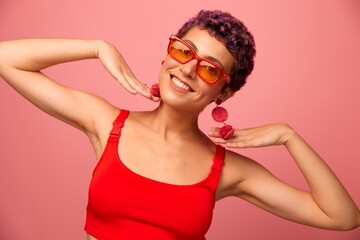 Young athletic woman with a short haircut and purple hair in a red top and pink yoga leggings in sunglasses with an athletic figure smiles and dances on a pink background