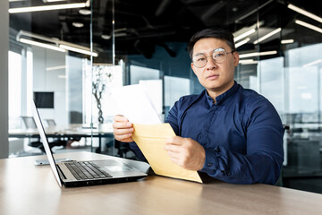 Fototapeta na wymiar Young Asian male lawyer in glasses sitting in the office at a desk with a laptop. He holds an envelope with documents in his hands. He looks seriously into the camera.