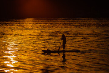 Naklejka na ściany i meble A lonely figure of a man floating on a board along the river. Sunset.