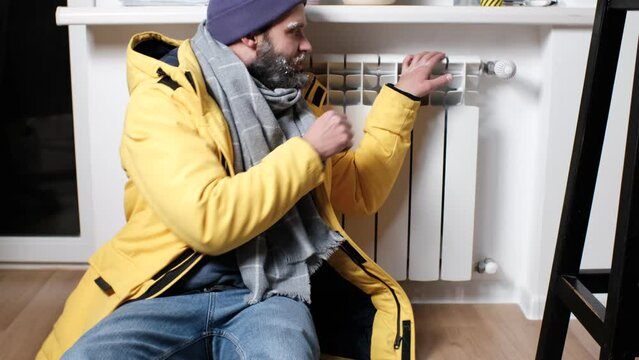 A frozen person is shivering from the cold in the apartment, a frozen face is covered with hoarfrost, in the hope of getting warm he touches a cold battery. The concept of the energy crisis in Europe